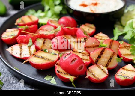 Grilled radishes with cucumber slices and sour cream sauce served on a dark grey plate, close-up, real photo, not AI generated Stock Photo