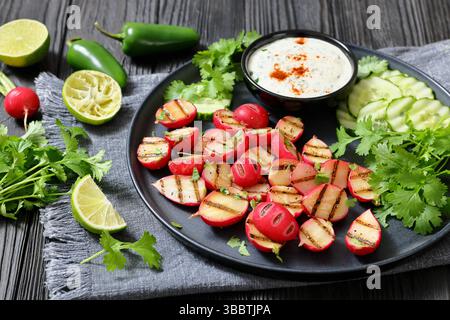 Grilled radishes with cucumber slices and sour cream sauce served on a dark grey plate on a black wooden table with fresh cilantro, jalapeno pepper an Stock Photo