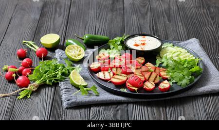 Grilled radishes with cucumber slices and sour cream sauce served on a dark grey plate on a black wooden table with fresh cilantro, jalapeno pepper, a Stock Photo