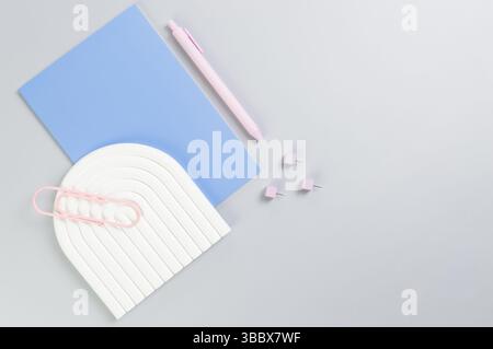 Top view of blue notebook, pink pen, paperclip, pins, white arch tray on grey background. School, office wallpaper. Flat lay, copy space. Stock Photo