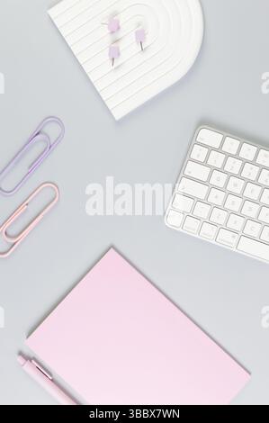 Top view of white keyboard, pink notebook, pink pen on grey background. Paper clips, pins, modern office, school. White arch tray, flat lay. Stock Photo