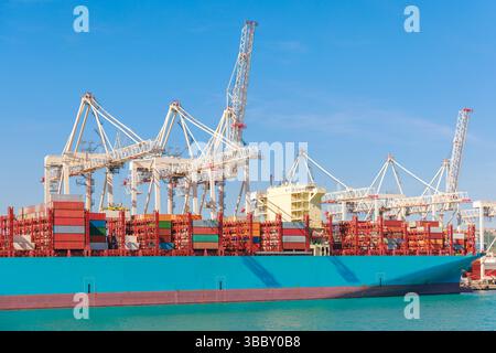 Large cargo ship is docked at a bustling shipping port, where cranes are actively loading containers for trade. Clear blue sky enhances the vibrant Stock Photo