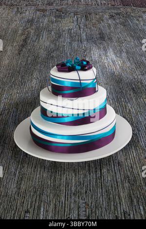 Minimalist still life image of a three tier hand baked cake decorated with blue and purple ribbons on a white porcelain plate placed on a wooden table Stock Photo