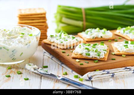 Crispy crackers and a glass bowl with a cream cheese spread and cut chives on a light kitchen table. Stock Photo