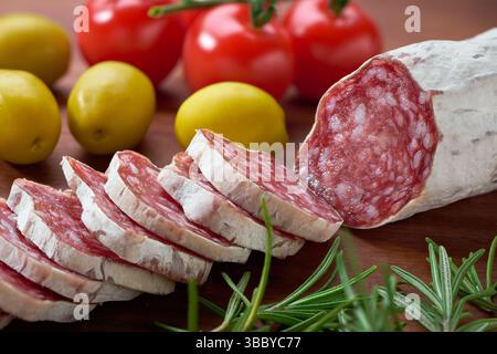 Traditional Spanish Fuet thin dried sausage with slices on a wooden board and next to olives and rosemary. Stock Photo