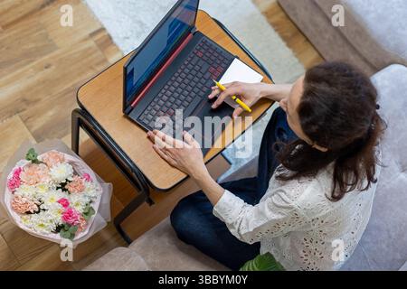 Woman working on laptop at home desk. Top view of a woman using a laptop and taking notes while sitting on a couch. Stock Photo