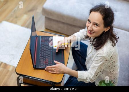 Smiling Woman Working on Laptop at Home. Happy woman sitting on sofa using laptop, holding pen and notepad, looking up and smiling in cozy home Stock Photo