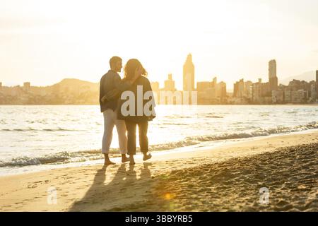 Young couple enjoying a peaceful beach stroll during golden hour, embracing and walking barefoot with the city skyline and mountains softly lit behind Stock Photo