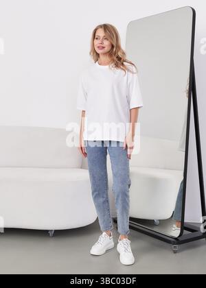 A woman in a white t-shirt and jeans stands in front of a large mirror, reflecting her image. She stands on a grey floor, beside a modern curved Stock Photo