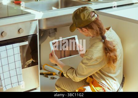 A woman plumber uses a tablet to inspect pipes under a kitchen sink, with plumbing tools scattered around her, indicating a repair. Stock Photo