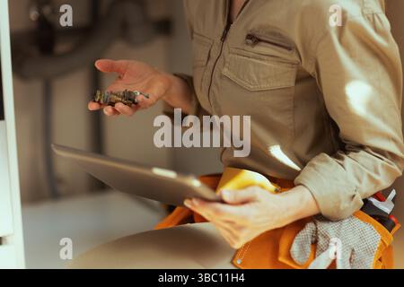 A fair-skinned plumber in a khaki jumpsuit uses a tablet to inspect an old plumbing valve fitting under a sink, likely troubleshooting a leak. Stock Photo