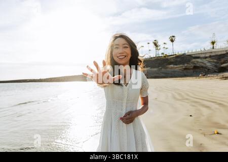 A woman is standing on a beach, smiling and with her open hand pointing at the camera, relaxed mood. Stock Photo
