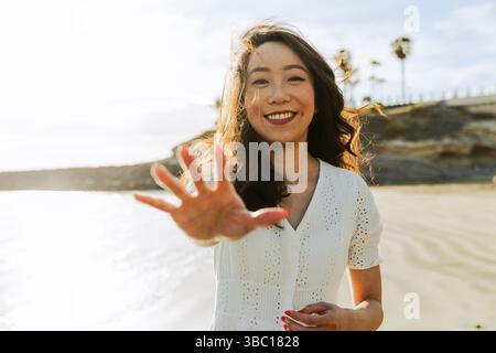 A woman is smiling and pointing her index finger at the camera. She is wearing a white shirt and is standing on a beach Stock Photo