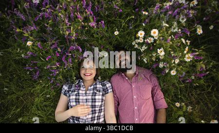 Young couple holding hands and lying outdoors in field with colorful wild flowers. Summer field with blooming wild peas and camomile flowers Stock Photo