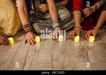 Women's hands holding lighted candles, sitting on floor. Indian holiday Diwali traditions. Closeup artificial candles on wooden floor. Nature saving c Stock Photo