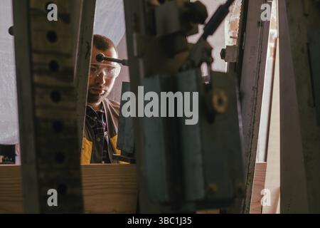 Closeup back view young professional woodworker in yellow vest, holding hydraulic press lever operating it to glue together pieces of wood. Woodworkin Stock Photo