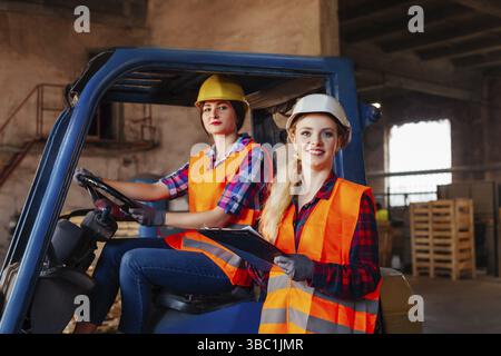 Young female forklift driver sitting in a truck talking to a woman delivery manager. Professional warehouse workers solving delivery issues. Cooperati Stock Photo