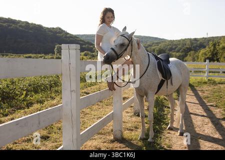 The young woman in a white dress is sitting on the fence and stroking a white horse on a ranch Stock Photo