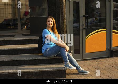 The happy female student in casual clothes is sitting on the stairs in front of the building Stock Photo