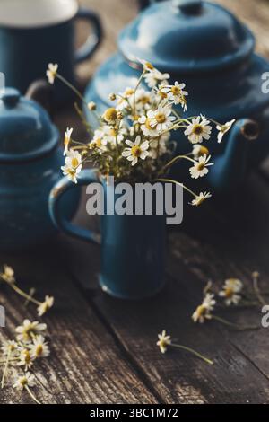 The bouquet of beautiful daisies in a mug on a wooden table. There is a blue enamel tea set nearby Stock Photo