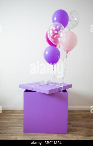The large purple gift box with balloons. The gift stands on the floor in the room on a background of a white wall Stock Photo