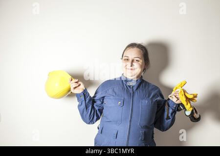 Smiling female worker dressed in work clothes, holding a yellow helmet and yellow gloves Stock Photo