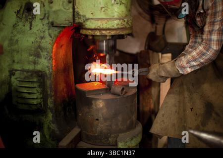 Blacksmith in safety gloves putting heated metal under press while working with metallurgical press machine. Worker using forging press in workshop Stock Photo
