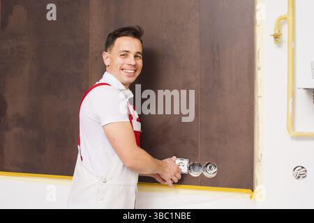 The smiling young man in overalls is fastening a new socket. The electrician works in the room where the repair takes place Stock Photo