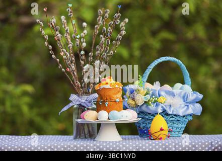 Spring sunny photo of Easter cake on a stand, surrounded by painted eggs, blue decorated basket, willow branches in glass. Green trees on a background Stock Photo