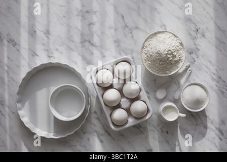 Top view ceramic tray with eggs placed near bowl of flour and sugar. White utensils with white ingredients on white surface. Copy space Stock Photo