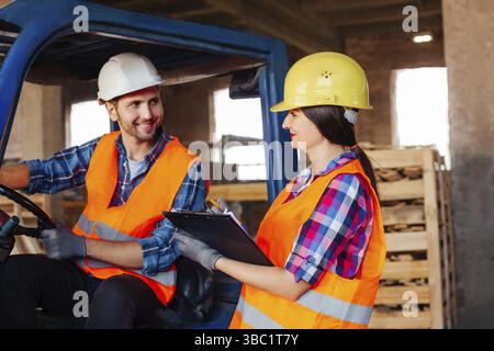 Male forklift driver sitting in a truck, female factory worker standing near, both discussing working issues. Warehouse workers in hardhats and orange Stock Photo