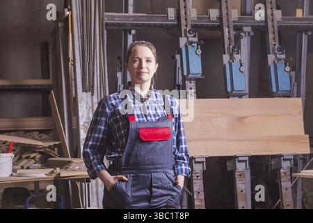 Portrait of attractive young female engineer at woodworking plant, wearing overalls, posing with hands in pockets. Woman feeling proud, working at car Stock Photo