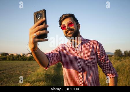 Portrait of young smiling man with stylish beard, wearing red sunglasses, taking selfie on summer field background. Outdoor photo of young confident l Stock Photo