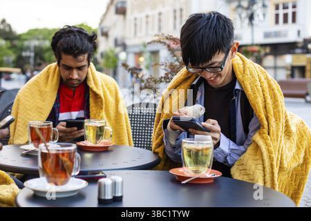 Three close diverse friends at the outdoor cafe, sitting covered with yellow plaids, watching their smartphones. Young men ordering food using online Stock Photo