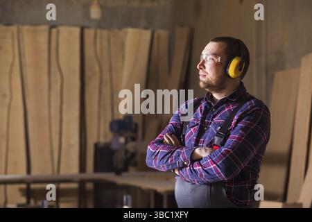 Portrait of the carpenter man in goggles and headphones. He stands in the factory and looks at the camera Stock Photo