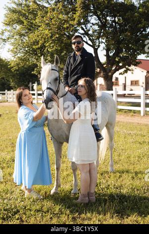 The young serious man in sunglasses and black jacket rides a white horse. The bearded man spends time on a horse farm Stock Photo