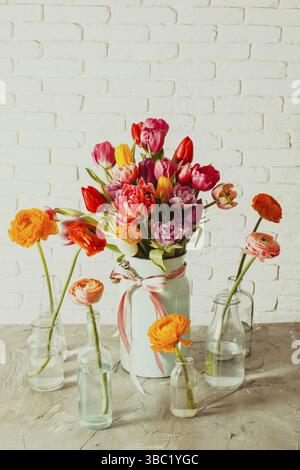 Six little glass bottles with ranunculus flowers inside standing around light metal vase filled with colorful fresh tulips. White wall with bricks tex Stock Photo