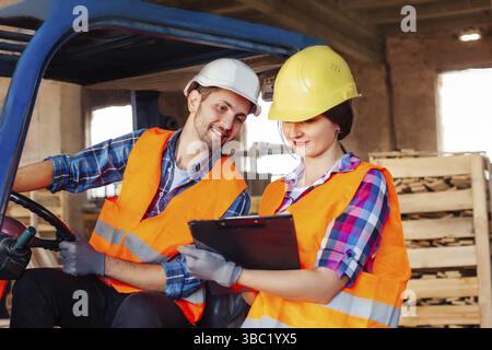 Male forklift driver sitting in a truck, female factory worker standing near, both discussing working issues. Warehouse workers in hardhats and orange Stock Photo