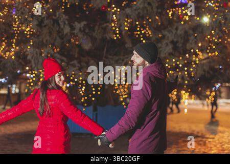 Backview portrait of young couple in bright colorful coats, standing holding hands near decorated fir tree on city square Stock Photo