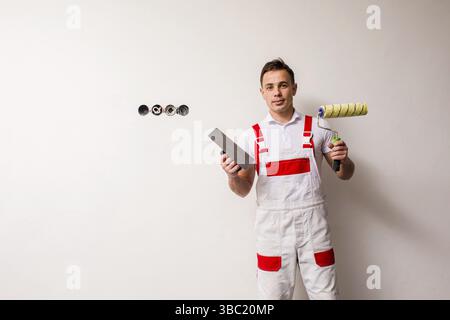Portrait of young smiling handyman with painting roller in one hand and trowel in another. Man in repair man's uniform on a white background, copy spa Stock Photo