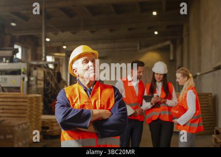 Happy aged man in protective working clothes and hard hat standing in front of group of young engineers at the factory. Experienced worker waiting for Stock Photo