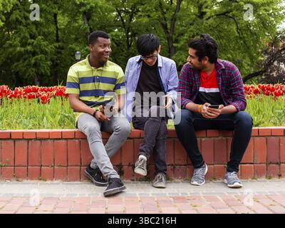 Three young close friends happy students meeting during break between lessons at college. Diverse male students sitting on edge of flower bed at city Stock Photo