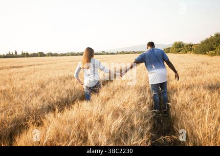 Cheerful couple holding hands while walking in grain field on warm sunny day. Diverse couple spending time far away from city rush Stock Photo