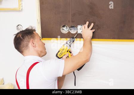 Male worker in overalls is fastening a new socket. The electrician works in the room where the repair takes place Stock Photo