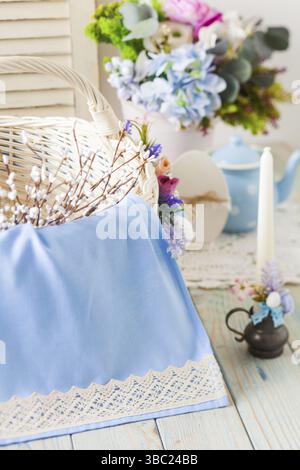 Easter concept. Light wicker basket full of willow branches. Close-up handmade blue cloth with crocheted white edging. Decorated candle and vase with Stock Photo