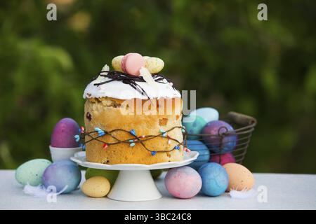 Photo of tasty Easter bread with original decoration of white icing, pink macarons and chocolate straw. Large amount of stylish, colored eggs near cak Stock Photo