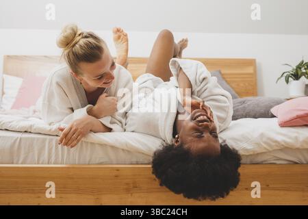 Two young women of different nationalities are lying on the bed at the spa and talking. The african woman is telling secret to her friend Stock Photo