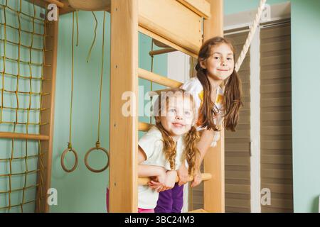 Two cute cheerful kids having fun on rope-ladder climbing and swingigng on it. Siblings strengthen themselves at home wooden sport complex Stock Photo