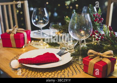 Christmas table setting with red crocheted Santa hat for cutlery on plates, festive decorations with fir tree branches. Christmas and New Year holiday Stock Photo