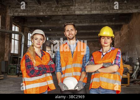 Two women and one man, young factory workers in orange protective vest and hard hats during work indoors old factory. Women standing with crossed hand Stock Photo
